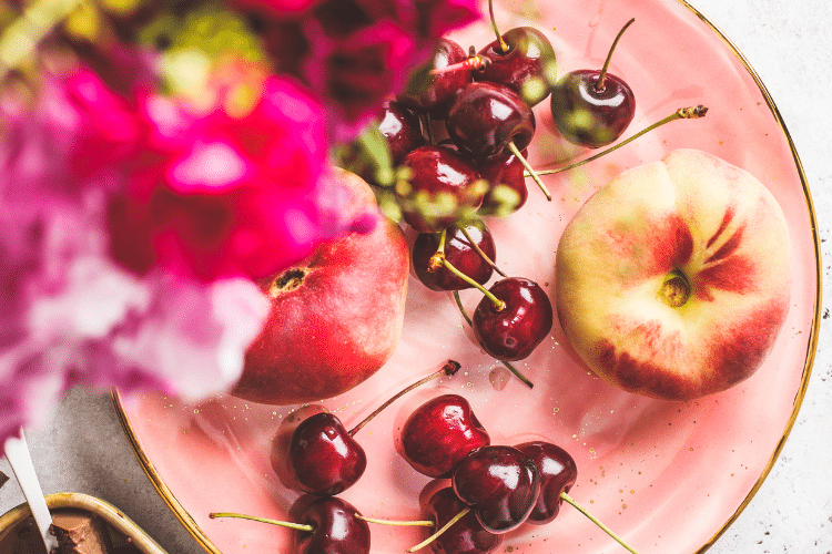 beautiful cherries and an apple arranged on a pink plate with pink flowers in the foreground