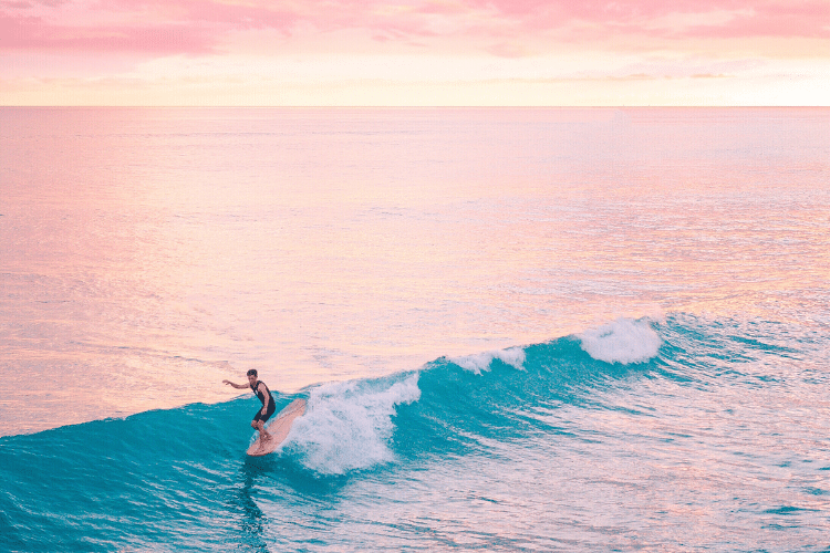 surfer riding a wave with pink clouds overhead creating a pink hue in the ocean
